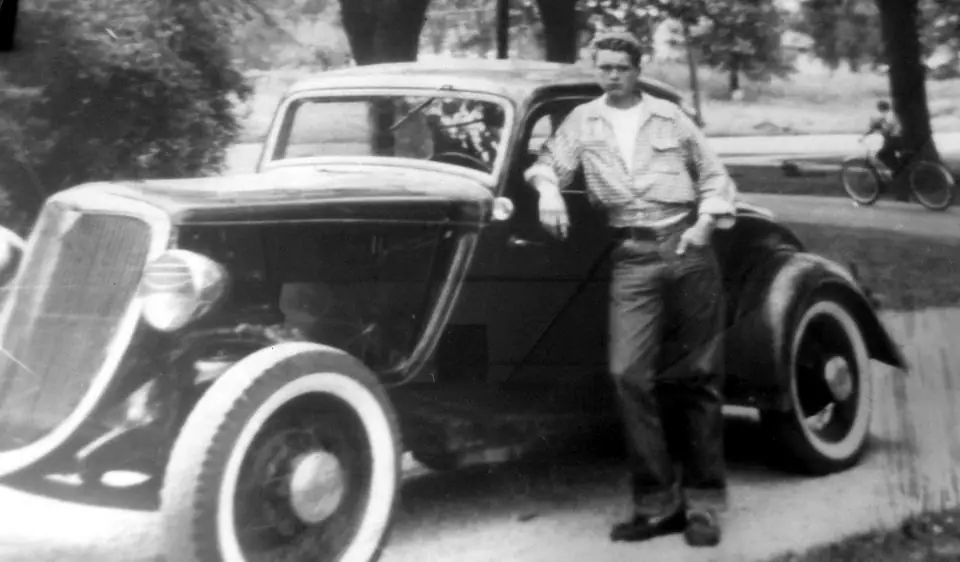 Black and white archive photo of a young man leaning on a 1930s coupe, from the Pfeiffer Auto Clinic website
